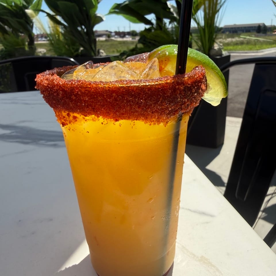 Large orange beverage with ice, lime wedge, straw, and chili-salt rim on an outdoor restaurant table.