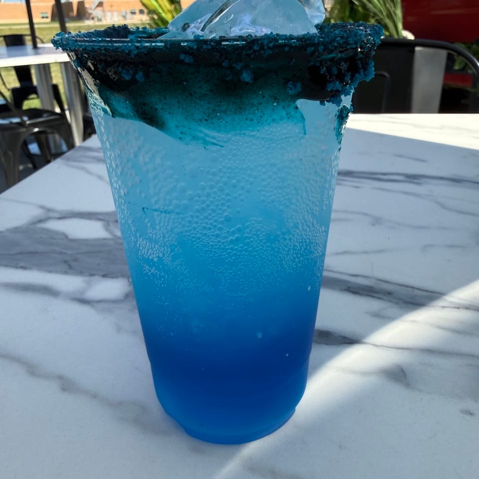 Bright blue cocktail with a blue sugar rim and ice, served on a marble patio table surrounded by greenery.