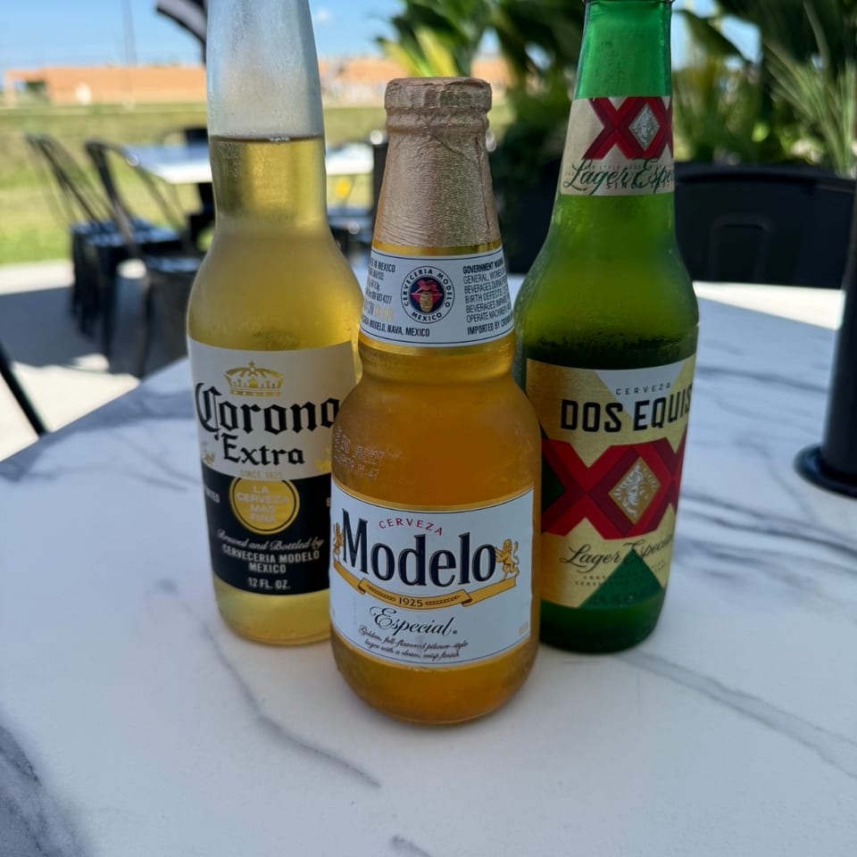 Three Mexican beer bottles—Corona Extra, Modelo Especial, and Dos Equis—sit on a patio table beneath umbrellas.