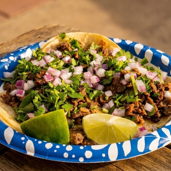 Three street tacos with meat, cilantro, and diced red onions, served with lime wedges on a paper plate.